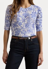 Kvinna i en tajt vit skjorta med blå blommönster, instoppad i mörkblå jeans med ett brunt bälte och silverfärgad spänne.