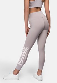 Leggings de color gris claro con una cinturilla alta, que presentan un gran logo blanco de "Lonsdale" en la pierna izquierda y una textura de tela lisa y elástica.