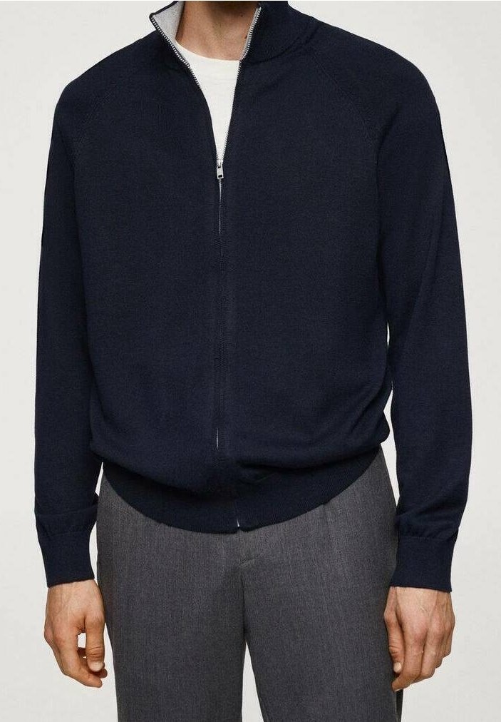 Homme portant un pull zippé bleu marine par-dessus une chemise blanche avec un pantalon gris, debout devant un fond blanc uni.