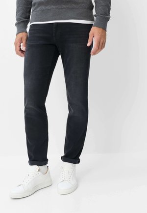 Man draagt donkere jeans met opgerolde pijpen, witte sokken, witte sneakers en een grijze trui, staand tegen een effen achtergrond.