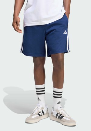 Mann trägt marineblaue Adidas-Shorts, weiße Adidas-Socken mit schwarzen Streifen und weiße Sneaker mit schwarzen Streifen, steht vor hellem Hintergrund.