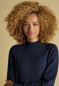 Dunkelblauer Pullover aus Strick mit einem gerippten Rundhalsausschnitt und langen Ärmeln. Glatte Textur, figurbetonter Schnitt. Die Person hat lockiges, blondes Haar.