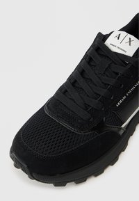 Detailný záber na čierne tenisky Armani Exchange s vrchnou časťou z mriežkovaného materiálu, zamakovanými akcentmi, šnúrkami a logom na jazyku a boku.