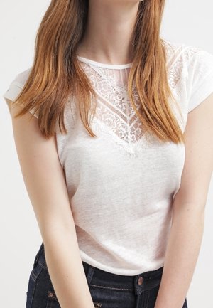 Mujer con una blusa blanca de mangas cortas con detalles de encaje, metida dentro de unos vaqueros azul oscuro, con cabello largo, liso y castaño claro visible.