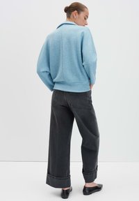 Vrouw met een haarstijl in een bun draagt een lichtblauwe jas, wijdvallende donkergrijze geclipte jeans en zwarte platte schoenen, staand tegen een neutrale studio-achtergrond.