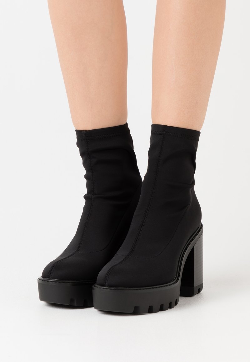 Jambes portant des bottines à talons hauts noires avec semelles plateformes épaisses sur fond blanc.