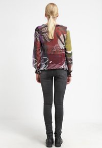 Blusa trasparente multicolore con motivi abstract e scritte; polsini aderenti; abbinata a jeans skinny neri e stivaletti.