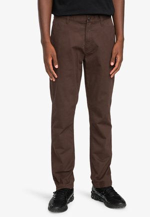 Pantaloni marroni a gamba dritta realizzati in twill di cotone. Presentano una chiusura anteriore con bottone, passanti per la cintura e tasche laterali. Indossati con scarpe nere.