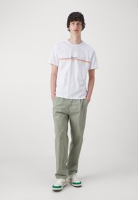 Fehér pamut póló színes vízszintes csíkokkal és "PEPE JEANS" logóval, világoszöld pamut nadrággal és zöld részletekkel ellátott fehér sportcipőkkel párosítva.