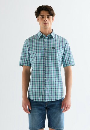 Młody mężczyzna w zielono-niebieską kratę na krótkim rękawie, zapinanej na guziki koszuli oraz niebieskich dżinsowych szortach na jasnym, jednolitym tle.