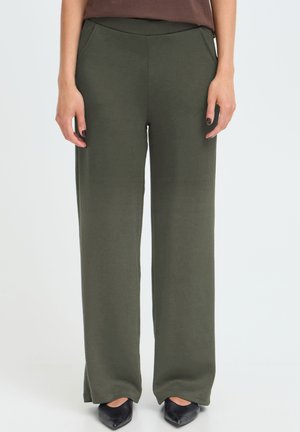 Femme portant un pantalon large vert olive avec poches avant et des ballerines noires pointues, debout devant un fond neutre.