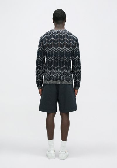 Πλεκτό πουλόβερ σε σκούρο μπλε με λευκό και γκρι ζιγκ ζαγκ μοτίβο, συνδυασμένο με σκούρα σορτς και λευκά αθλητικά παπούτσια. Πλάγια όψη από πίσω.