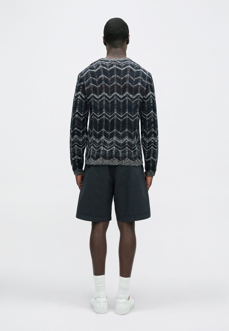 Pull-over tricoté bleu foncé avec motif en zigzag blanc et gris, assorti à un short foncé et des baskets blanches. Vue de dos.