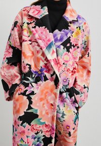 Kabát s květinovým vzorem na černém pozadí, vyrobený z jemné texturované látky, s velkými knoflíky a upraveným límcem.