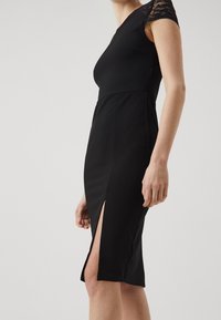 Schwarzes Kleid mit eng anliegendem Oberteil und seitlichem Schlitz. Verfügt über Spitzenverzierungen an den kurzen Ärmeln, eine glatte Textur und einen knielangen Schnitt.