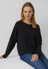 Zwarte sweater met lange mouwen, een losse pasvorm, ronde hals en raglanmouwen. Gecombineerd met blauwe spijkerbroek, tegen een neutrale achtergrond.