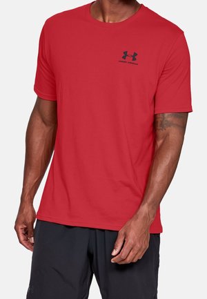 Man draagt een rood Under Armour T-shirt met korte mouwen en een zwarte short, zichtbaar van schouders tot midden dijen, met een tatoeage op de linkerarm.
