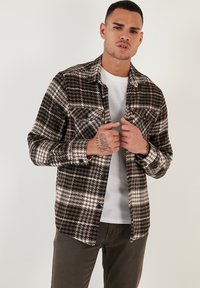 Cămașă flanel cu carouri maro și alb, cu design cu nasturi și două buzunare pe piept, purtată cu un tricou alb și pantaloni gri.