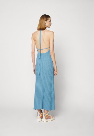 Femme aux cheveux courts bruns portant une robe maxi en tricot bleu sans dos et des baskets montantes blanches ornées de pompons colorés, se tenant devant un fond blanc.