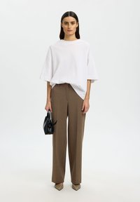 Femme aux longs cheveux bruns portant un t-shirt blanc ample, un pantalon marron à jambes larges, tenant un petit sac à main noir, debout sur un fond blanc.