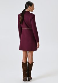Cappotto bordeaux con maniche lunghe, un taglio aderente e una vita dettagliata. Stivali da cowboy marroni ricamati completano l'outfit.