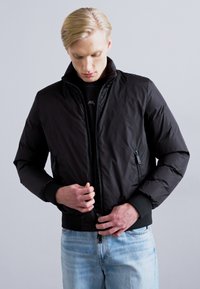 Black puffer jacket with a high collar, front zipper, side pockets, and ribbed cuffs. Worn over a black shirt and light blue jeans.