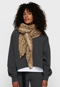 Beige tørklæde med et gentaget sort logo mønster, løst draperet om halsen over en mørkegrå sweatshirt. Blød stofstruktur, letvægtsdesign.