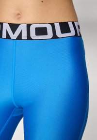 Modre športne legice z gladko teksturo in črnim pasom z belimi črkami. Prikaz podrobnosti oblikovanja v bližnjem planu.