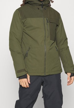 Mann trägt grüne Kapuzenjacke mit Reißverschlusstaschen an Brust und Seiten, kombiniert mit schwarzer Hose, steht vor weißem Hintergrund.