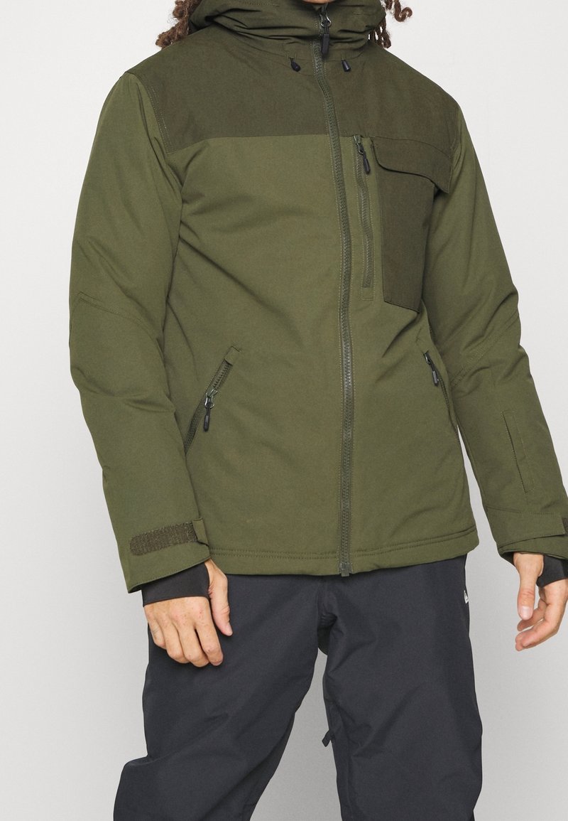 Homme portant une veste à capuche verte avec poches zippées sur la poitrine et les côtés, assortie à un pantalon noir, debout devant un fond blanc.