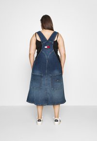 Džínová šaty, tmavě modrá s dvěma předními kapsami, nastavitelnými ramínky a značkovým štítkem na zadní straně. Měkká textura, délka po lýtka.