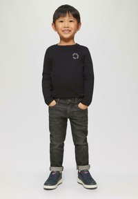 Lachende jonge jongen staat met zijn handen in zijn zakken, gekleed in een zwart langarmshirt, donkere opgerolde jeans en marineblauwe sneakers, tegen een effen achtergrond.