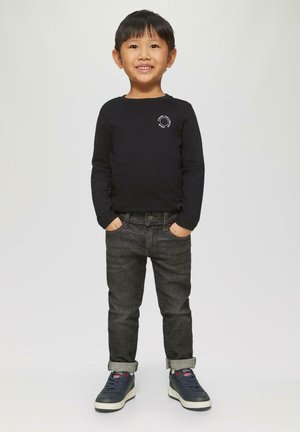 Lachende jonge jongen staat met zijn handen in zijn zakken, gekleed in een zwart langarmshirt, donkere opgerolde jeans en marineblauwe sneakers, tegen een effen achtergrond.