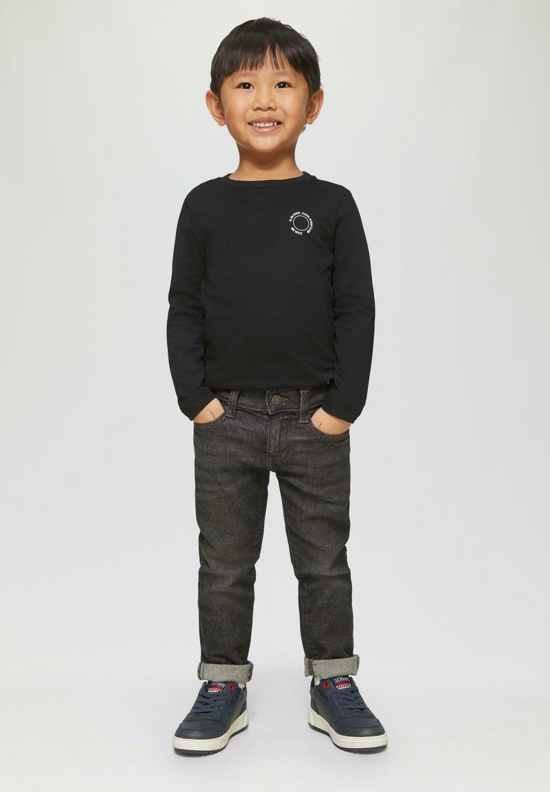 Lachende jonge jongen staat met zijn handen in zijn zakken, gekleed in een zwart langarmshirt, donkere opgerolde jeans en marineblauwe sneakers, tegen een effen achtergrond.