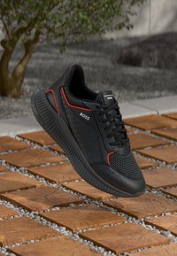 Black athletic shoe featuring a perforated upper, red accents, and a textured rubber sole, set against a tiled ground with pebbles.