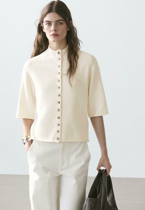 Vrouw met lang bruin haar draagt een crèmekleurig kort mouwen topje met knopen en een witte broek, houdt een grote donkerbruine leren tas in één hand.