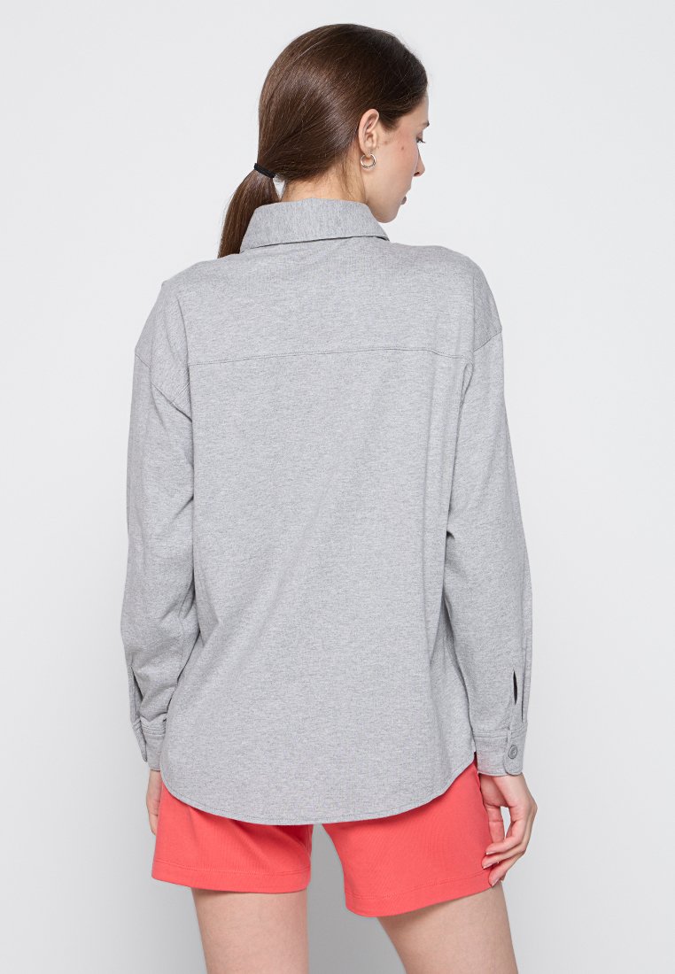 Graues Langarmhemd mit Kragen, Rückenpasse und geknöpften Manschetten, kombiniert mit korallenfarbenen Shorts. Schlichte Stofftextur.