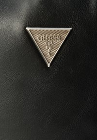 Čierny textúrovaný materiál s trojuholníkovým strieborným emblémom s nápisom "GUESS" a detailom otáznik. Jednoduchý a geometrický dizajn.