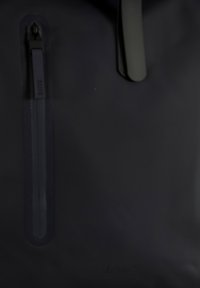 Αδιάβροχη μαύρη τσάντα με έντονη τσέπη με φερμουάρ, λεία υφή, μινιμαλιστικό σχέδιο και ανάγλυφο λογότυπο της μάρκας.