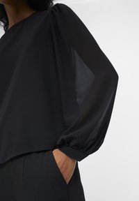Blusa negra con mangas largas de tul y una silueta relajada. Material suave con un brillo sutil, que presenta un escote redondeado.