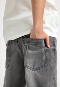 Grå denimjeans med en blekt finish, som har två bakfickor. Subjektets vita skjorta är delvis synlig, med ett strukturerat tyg.