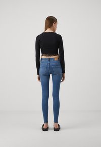 Crna majica s dugim rukavima i čipkastim rubom, uparena s plavim skinny trapericama. Model prikazan s leđa s glatkom smeškom smeškom i crnim cipelama.