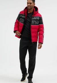 Rote gep padded Jacke mit schwarzen Akzenten, ausgestattet mit auffälligen Aufdrucken und Reißverschlusstaschen, kombiniert mit schwarzen Hosen und Turnschuhen. Mit angebrachtem Kapuzen. 