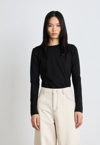 Black long-sleeve t-shirt with a round neckline and a small blue logo, paired with cream-colored high-waisted wide-leg pants with contrast seams.