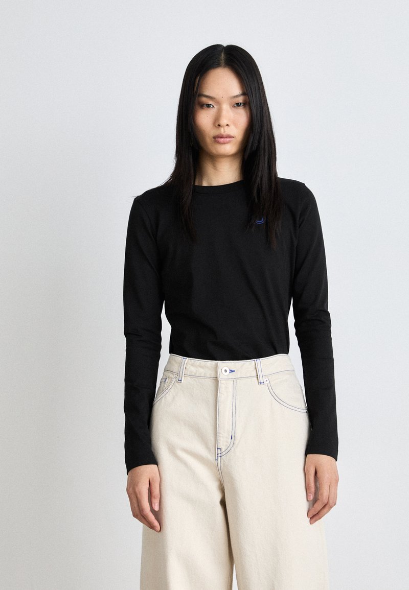 Black long-sleeve t-shirt with a round neckline and a small blue logo, paired with cream-colored high-waisted wide-leg pants with contrast seams.