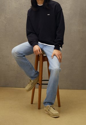 Suéter negro con el logo "LEVI'S", jeans desgastados de color azul claro y zapatos de ante color beige. Modelo sentado en un taburete de madera contra un fondo gris.