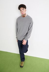 Gray crew-neck sweater, navy pants, and camo hiking boots. Casual fit with ribbed cuffs. Standing on green carpet against a white wall.