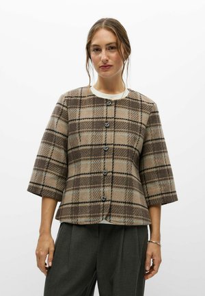 Une femme porte une veste à carreaux beige et marron, aux manches trois-quarts et ornée de boutons décoratifs, assortie à un pantalon foncé sur fond blanc.