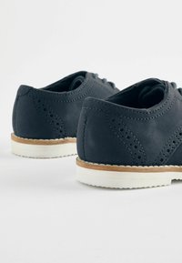Chaussures en cuir bleu foncé avec des détails perforés, semelles en caoutchouc blanches et accents crème sur le bas. Forme arrondie avec lacets.