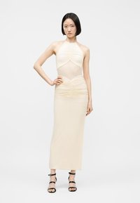 Γυναίκα όρθια με το χέρι στη μέση, φορώντας αμάνικο κοντό φόρεμα σε κρεμ χρώμα με ρυτίδες και μαύρα ψηλοτάκουνα σανδάλια με λουράκια, σε λευκό φόντο.